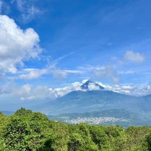 Un gran volcán se alza sobre un pueblo en el valle, visto sobre un exuberante bosque verde bajo un cielo azul parcialmente nublado.