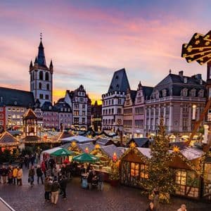 Un vivace mercatino di Natale riempie una piazza lastricata di ciottoli con bancarelle illuminate e una grande piramide di legno sotto un cielo al tramonto.