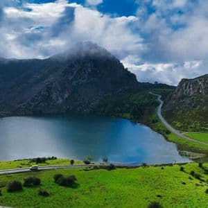 Una vista aerea di una strada tortuosa lungo un lago calmo, incastonata tra verdi colline e montagne sotto un cielo nuvoloso.