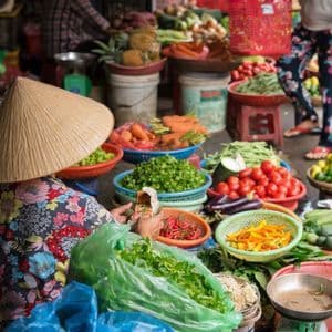 Vu de dos, un vendeur portant un chapeau conique est assis entouré de paniers de légumes frais sur un étal de marché en plein air.
