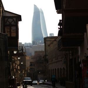 Un moderno grattacielo di vetro si erge su una stretta strada acciottolata fiancheggiata da edifici tradizionali in ombra.