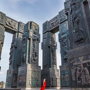 Una donna con un lungo abito rosso si erge davanti a colossali monumenti in pietra scolpita in un'ampia piazza aperta.