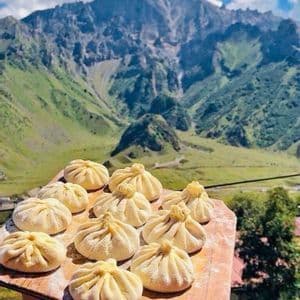 Ravioli pieghettati crudi disposti su un tagliere di legno, sullo sfondo di montagne verdi.