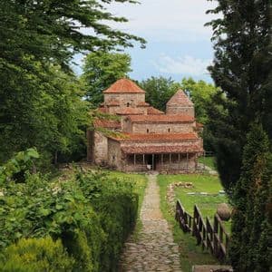 Un rustico monastero in pietra con tetti di tegole rosse sorge alla fine di un sentiero acciottolato, immerso in una lussureggiante vegetazione verde.