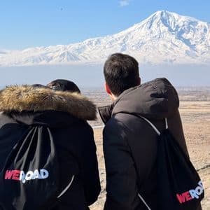 Due persone di un viaggio di gruppo WeRoad, viste di spalle, che guardano una grande montagna innevata sotto un cielo azzurro e limpido.