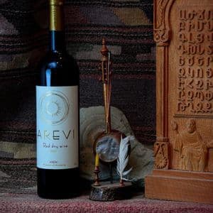 Una bottiglia di vino rosso armeno è disposta accanto a un minuscolo strumento a corda e a una targa in legno intagliato su un tessuto testurizzato.