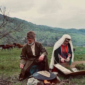 Un uomo e una donna in abiti tradizionali cucinano pane piatto su un falò in un prato collinare, con cavalli sullo sfondo.