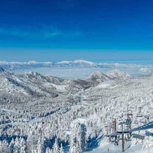 Una vista panoramica da una seggiovia mostra una vasta catena di montagne e foreste innevate sotto un cielo azzurro brillante.