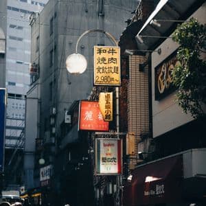 Eine Menschenmenge geht eine schmale Stadtstraße entlang, die von Gebäuden und vertikalen Schildern mit japanischen Schriftzeichen gesäumt ist.