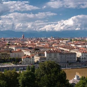 Una vista panorámica de una ciudad con tejados de terracota y una aguja prominente, con montañas a lo lejos bajo un cielo azul nublado.