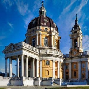 Una gran basílica barroca con fachada amarilla, cúpula oscura y campanario contra un cielo azul vibrante.