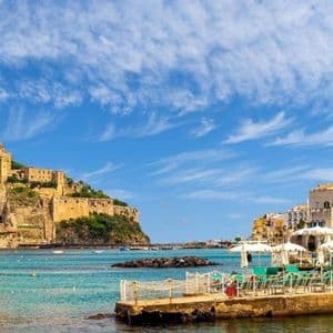 Un castello si erge su una scogliera rocciosa che domina una città costiera e una baia piena di barche sotto un cielo azzurro.