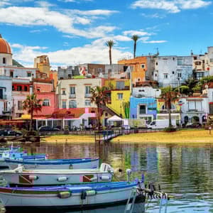 Piccole imbarcazioni sono ormeggiate in un porto di fronte a una colorata città costiera con una spiaggia sabbiosa e una chiesa a cupola.