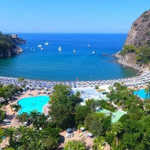 Vista aerea di un resort costiero con piscine e una spiaggia sabbiosa che costeggia una baia con barche sull'acqua blu.