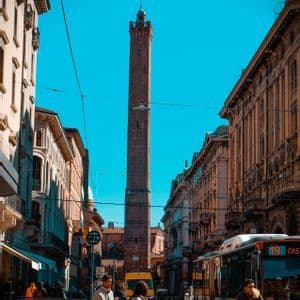 Una alta torre de ladrillo se alza al final de una estrecha calle de la ciudad con gente, bicicletas y un autobús bajo un cielo azul claro.