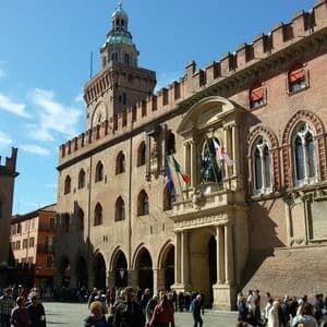 Una multitud de personas se reúne en una plaza histórica de la ciudad frente a un gran edificio de ladrillo rojo con una torre del reloj bajo un cielo azul.