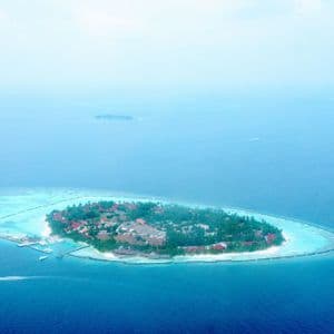 Vue aérienne d'un petit complexe hôtelier tropical sur une île, avec des palmiers, entouré d'un lagon turquoise et d'un océan bleu profond.