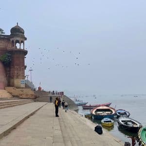 Des ghâts en pierre descendent vers une rivière où sont amarrés des bateaux colorés, avec des gens qui passent par un jour de brouillard.