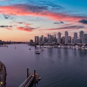 Eine Stadtsilhouette mit modernen Wolkenkratzern überblickt bei Sonnenuntergang einen Hafen mit Booten unter einem farbenfrohen bewölkten Himmel.