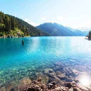 Ein klarer türkisfarbener Alpensee mit felsigem Ufer im Vordergrund, umgeben von mit Kiefern bewachsenen Bergen.