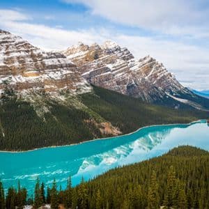 Ein leuchtend türkiser See spiegelt schneebedeckte Berge wider, umgeben von dichten Kiefernwäldern unter einem teilweise bewölkten Himmel.