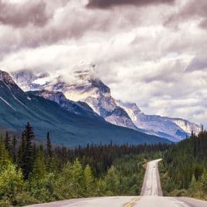 Eine asphaltierte Straße führt durch einen dichten immergrünen Wald zu einem großen, felsigen Gebirge unter einem bewölkten Himmel.