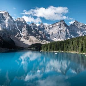 Ein türkiser Alpensee spiegelt eine felsige Bergkette und bewölkten blauen Himmel wider, mit einem dichten Kiefernwald am Ufer.