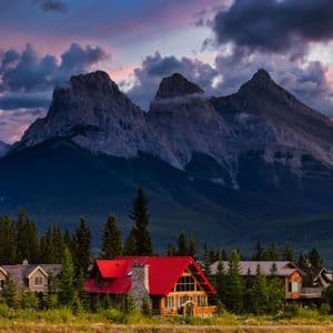 Holzhäuser, eines mit leuchtend rotem Dach, eingebettet in einem Kiefernwald am Fuße zerklüfteter Berge unter einem bewölkten Sonnenuntergang.