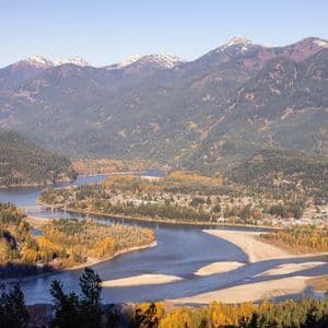 Luftaufnahme eines breiten Flusses mit Sandbänken, der durch ein Herbsttal fließt, mit einer kleinen Stadt und schneebedeckten Bergen.