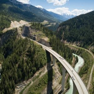 Luftaufnahme einer Autobahnbrücke mit Autos, die sich über ein bewaldetes Tal mit einem Fluss und Bergen schlängelt.