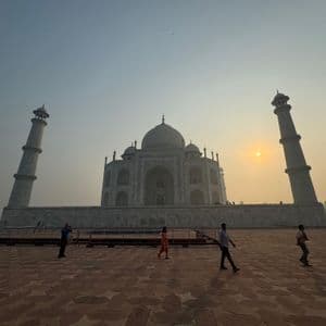 Gente camina por un patio de piedra frente a un gran mausoleo de mármol blanco flanqueado por minaretes al atardecer.