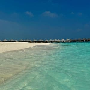 Agua cristalina turquesa baña una playa de arena blanca bordeada de bungalows sobre el agua bajo un cielo azul profundo.
