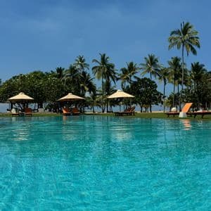 Una gran piscina en un resort tropical con tumbonas y sombrillas en un césped bordeado de palmeras bajo un cielo azul claro.