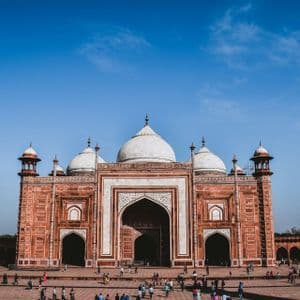 Un edificio ornamentado de arenisca roja con cúpulas blancas se alza sobre un gran patio lleno de gente bajo un cielo azul brillante.