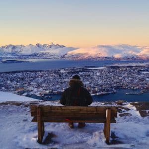 A person sits on a bench on a snowy mountaintop, overlooking a coastal city and distant mountains at sunset.
