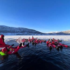 A WeRoad group trip wearing red immersion suits floating in a frozen lake, surrounded by a snowy landscape and mountains.