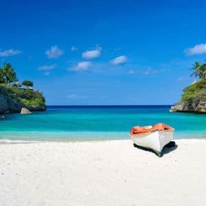 Una piccola barca con interni arancioni riposa su una spiaggia di sabbia bianca in una cala con acqua turchese e scogliere coperte di vegetazione.