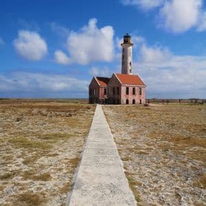 Un lungo sentiero di cemento conduce a un faro logoro e a una costruzione annessa in un paesaggio piatto e arido sotto un cielo azzurro.