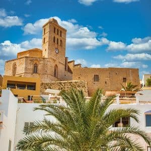 Una iglesia de piedra con un campanario se alza en una colina sobre edificios blancos, con una gran palmera en primer plano bajo un cielo azul.
