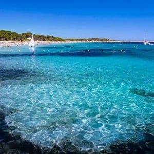 Agua turquesa cristalina ondea en primer plano de una playa soleada con veleros y gente en la arena bajo un cielo azul.