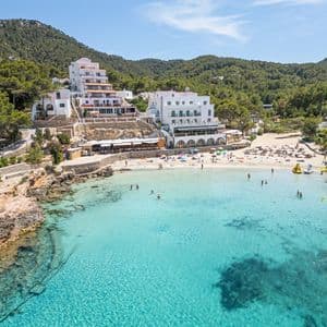 Vista aérea de una cala de playa con gente nadando en agua turquesa y tomando el sol en la arena, respaldada por hoteles blancos en una ladera verde y arbolada.