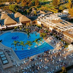 Vista aerea di un grande resort al tramonto, con piscina affollata, palme e edifici moderni.