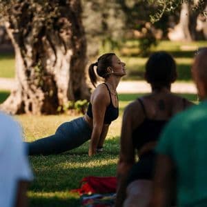 Una donna con auricolare guida un gruppo WeRoad durante una lezione di yoga all'aperto su un prato erboso, sotto gli alberi.
