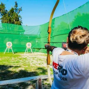 Una capogruppo WeRoad con i capelli biondi raccolti in uno chignon mira con arco e freccia a dei bersagli su un campo di tiro con l'arco all'aperto.