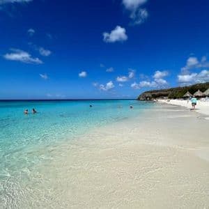 Persone che nuotano in acqua cristallina turchese su una spiaggia di sabbia bianca sotto un cielo blu intenso con nuvole bianche.