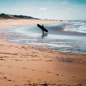 Un surfista in silhouette porta una tavola da surf lungo la riva di una spiaggia sabbiosa mentre le onde si infrangono.