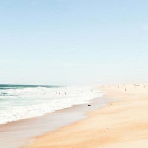 Le onde dell'oceano lambiscono una vasta spiaggia sabbiosa, dove le persone nuotano e si rilassano sotto un cielo azzurro pallido.