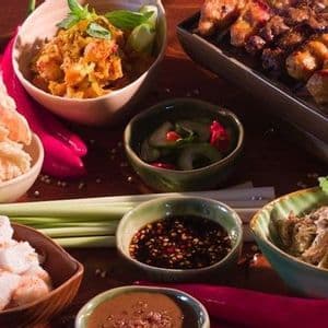 Un assortiment de divers plats asiatiques, incluant des brochettes de satay, des crackers, des salades et des sauces pour tremper sur une surface en bois.