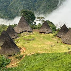 Un village traditionnel avec des huttes coniques aux toits de chaume sur une colline verdoyante, entouré de montagnes boisées et de nuages bas.