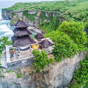 Une vue aérienne d'un temple traditionnel aux toits de chaume, perché sur une falaise verdoyante dominant l'océan.